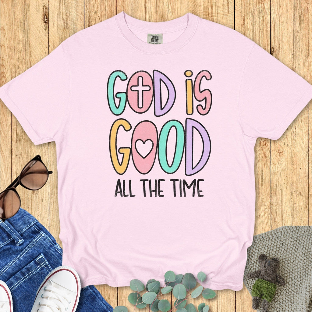 Flatlay of a Comfort Colors 1717 blossom pink “God Is Good All The Time” t-shirt in cute pastel lettering with cross and heart icons. A sweet women’s scripture tee -  faith-based apparel for women and modern Christian shirt, ideal Christian mom gift idea for daily faith wear.