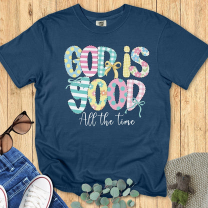 Flatlay of a Comfort Colors 1717 midnight “God Is Good All the Time” t-shirt featuring playful patchwork typography and bow details. A cheerful women’s scripture tee — faith-based apparel for women and modern Christian shirt, perfect Christian mom gift idea.