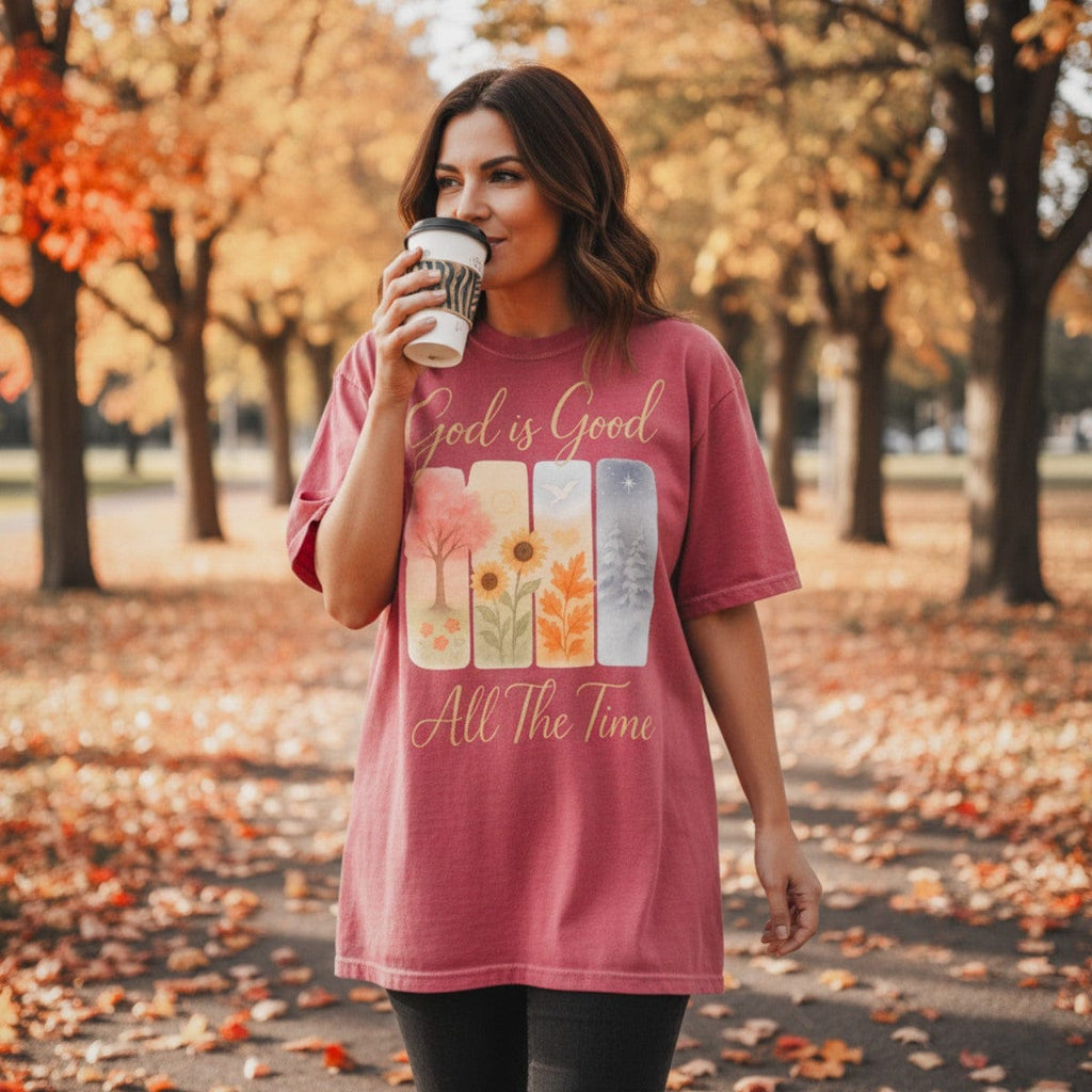 Christian women wearing a Comfort Colors 1717 crimson t-shirt with the inspirational design "God is Good All The Time." The seasonal artwork features spring, summer, fall, and winter scenes, captured in an autumn park setting with fallen leaves as she holds a coffee cup.