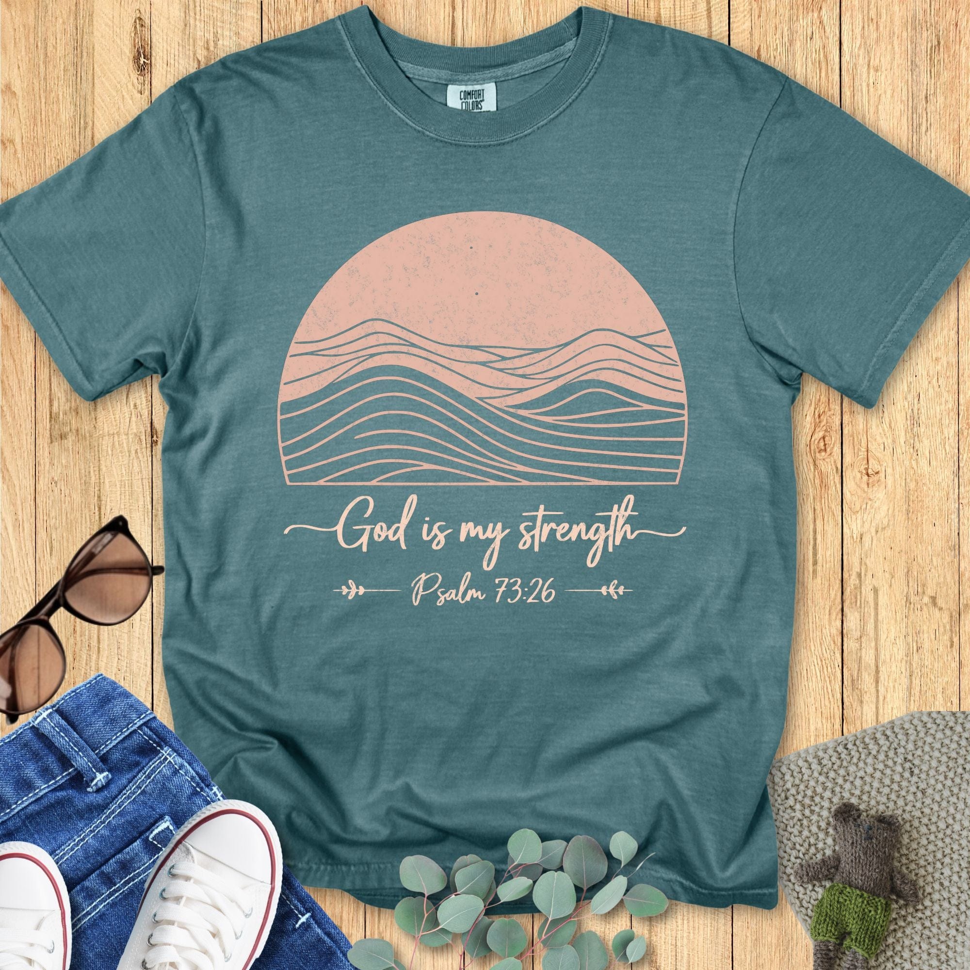 Blue spruce Comfort Colors 1717 Christian t-shirt with pink sun and wave graphic and the text 'God is my strength – Psalm 73:26,' arranged flat lay with denim, sneakers, sunglasses, and greenery accents