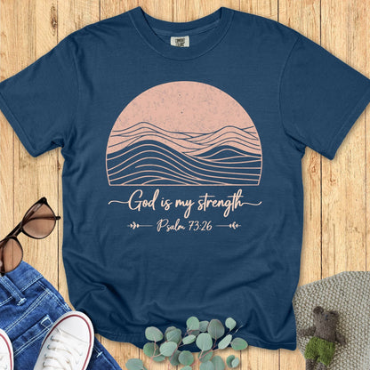 Midnight blue Comfort Colors 1717 Christian t-shirt with pink sun and wave illustration and the words 'God is my strength – Psalm 73:26,' styled with jeans, sneakers, sunglasses, and eucalyptus leaves on wood background