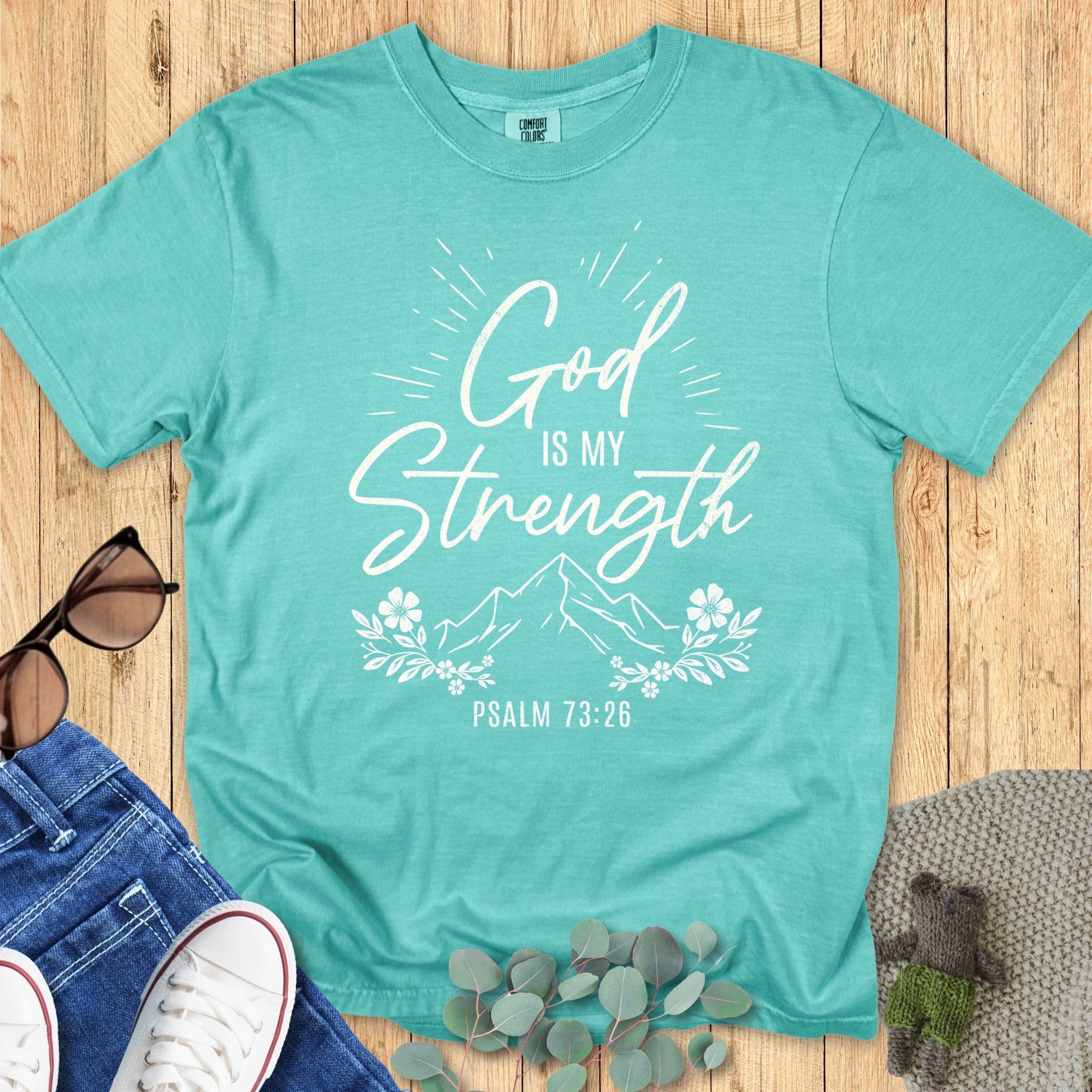 Comfort Colors 1717 seafoam green unisex shirt with white Christian design “God is my Strength – Psalm 73:26.” Features mountains, florals, and radiating sun rays. Flat lay with jeans and casual props