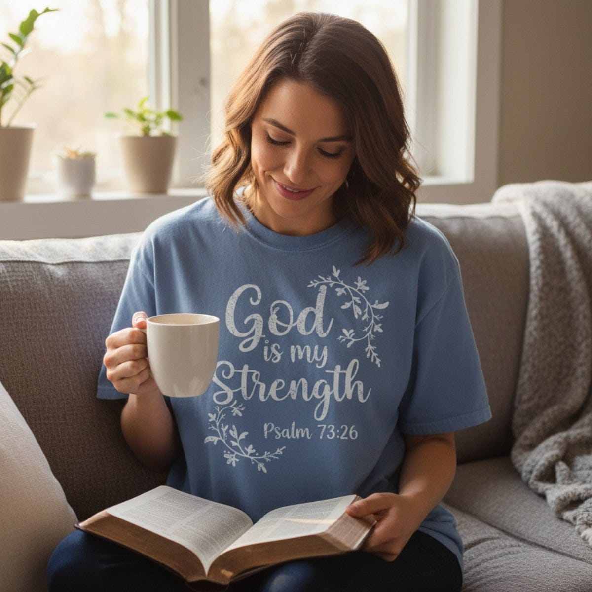 Woman sitting on a sofa, wearing a blue Comfort Colors 1717 Christian apparel T-shirt with the words "God is my Strength, Psalm 73:26" in white floral script, holding a coffee mug and reading a Bible.