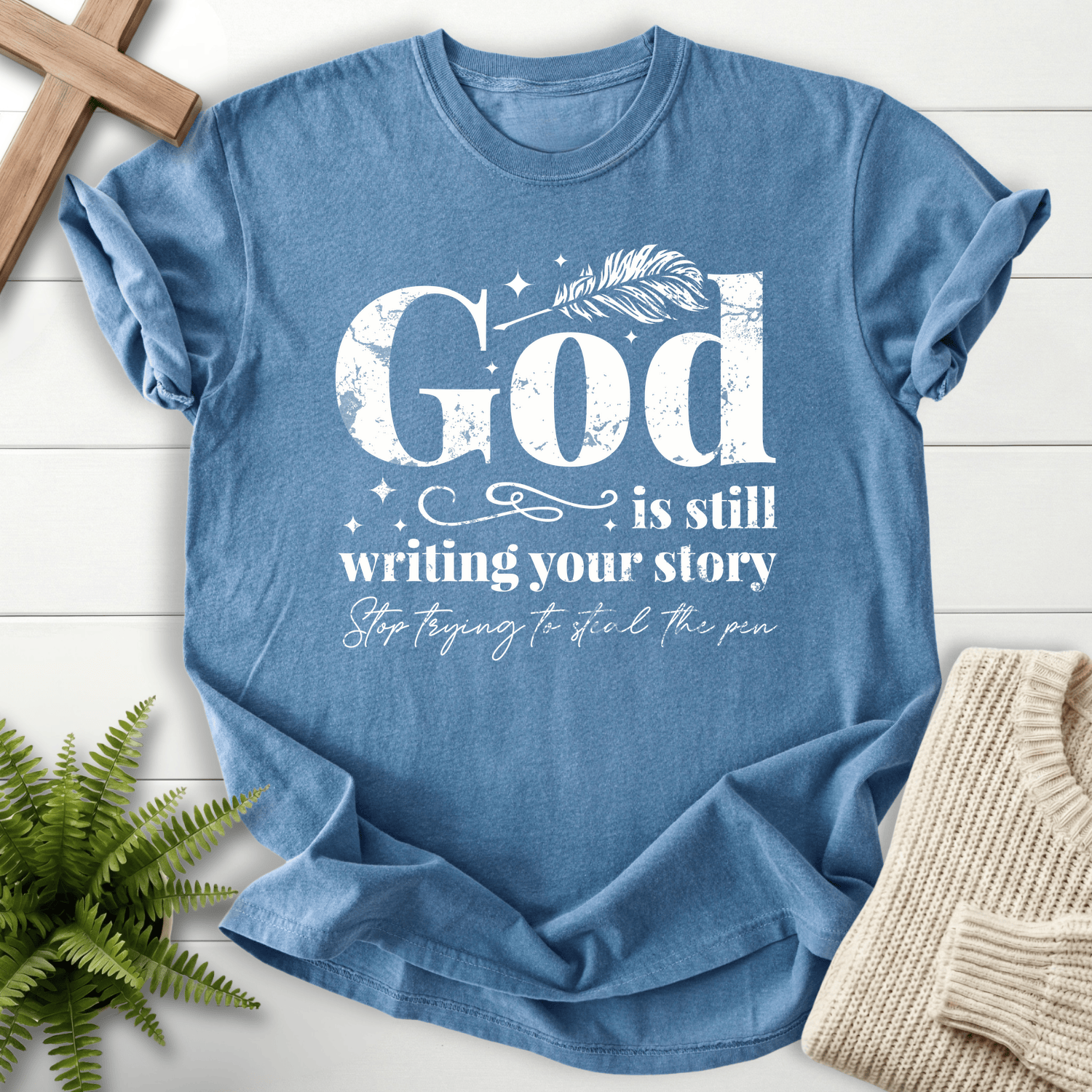 Blue Jean women's faith t-shirt with grunge 'God is Still Writing Your Story' lettering and a quill pen motif.