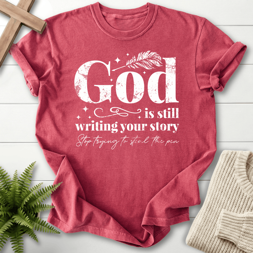 Crimson women's faith t-shirt with a feather quill and bold 'God is Still Writing Your Story' in grunge font.