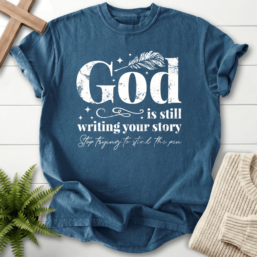Midnight blue Christian t-shirt for women with distressed white 'God is Still Writing Your Story' and a quill pen accent.