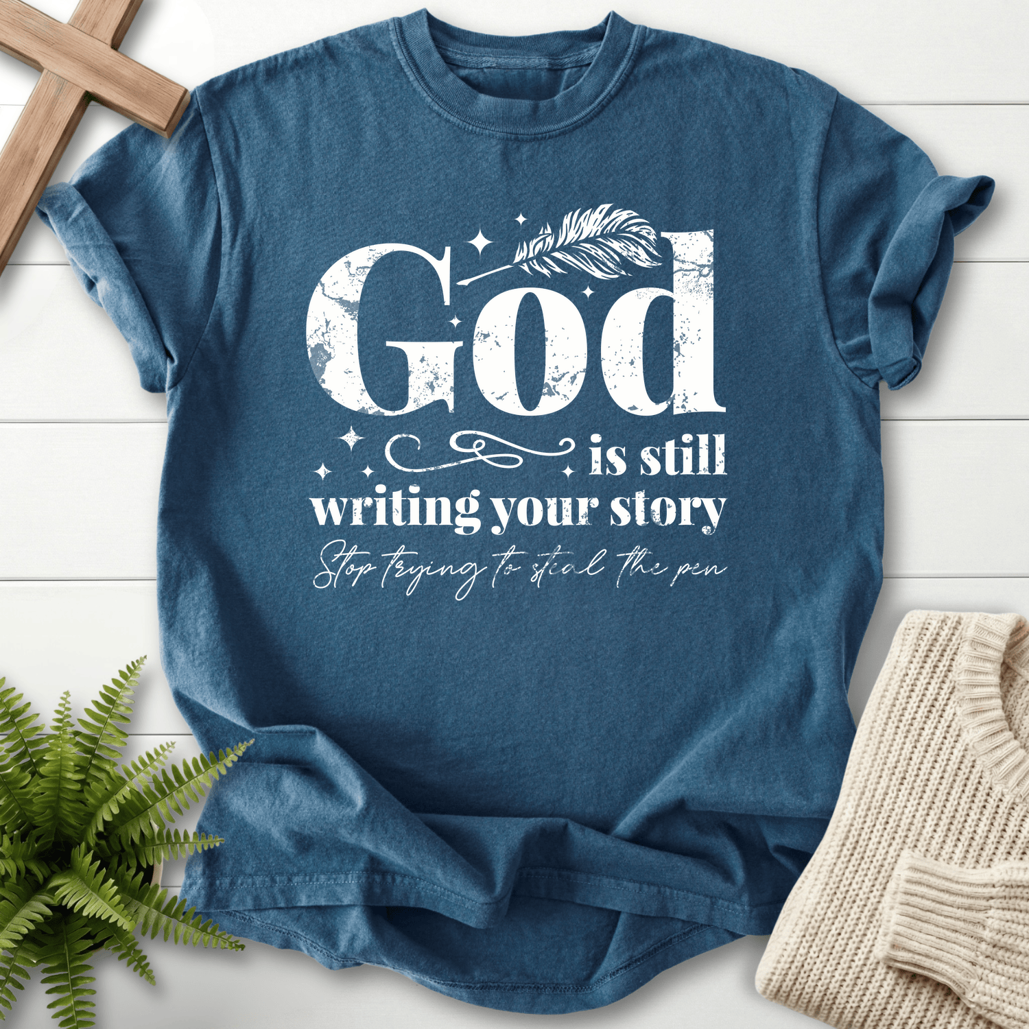 Midnight blue Christian t-shirt for women with distressed white 'God is Still Writing Your Story' and a quill pen accent.