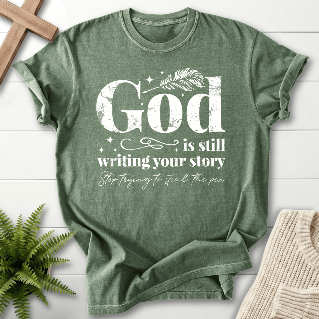 Moss green women's faith t-shirt with 'God is Still Writing Your Story' in grunge lettering and the phrase 'Stop trying to steal the pen'.