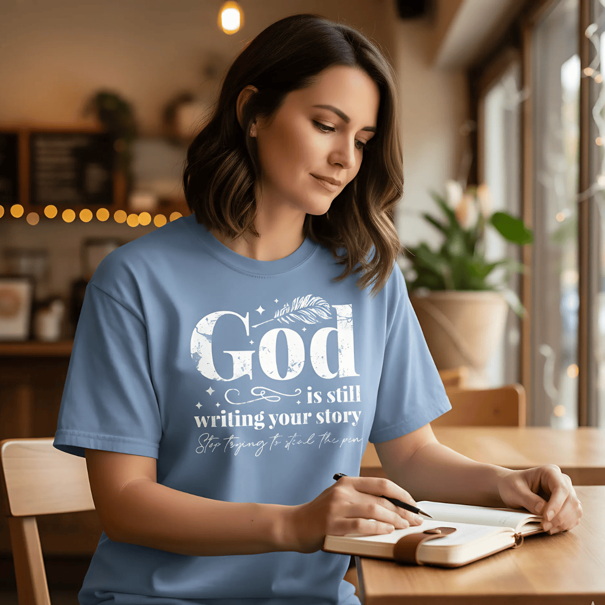 Woman journaling at a cozy café wearing a blue jean Comfort Colors 1717 Christian T-shirt with white design reading “God is still writing your story, stop trying to steal the pen