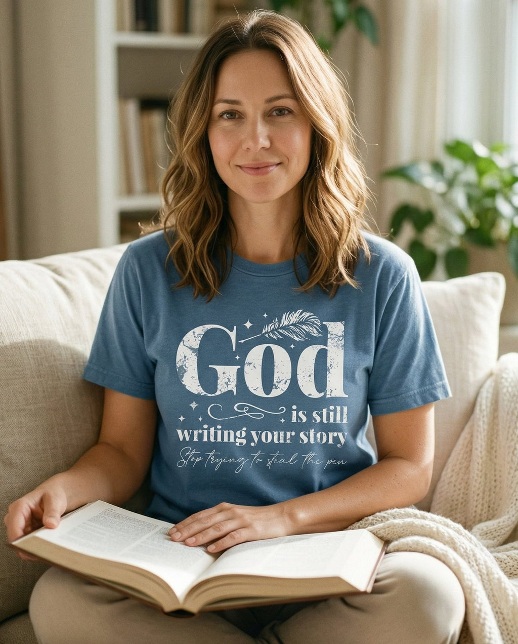 God is Still Writing Your Story T-shirt – Blue Jean women's Christian tee, lifestyle photo in cozy living room – Distressed grunge-style 'God is still writing your story' with a feath – She Wears Faith