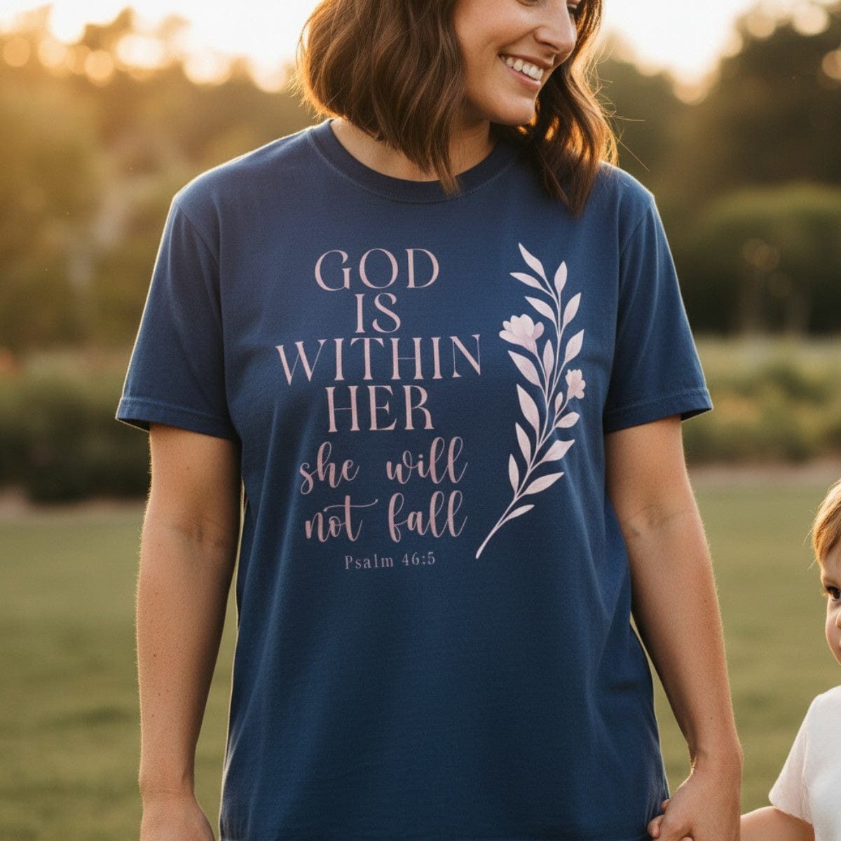 Smiling woman outdoors at sunset wearing a Comfort Colors 1717 midnight “God Is Within Her – Psalm 46:5” t-shirt with soft pink floral design. A meaningful women’s scripture tee and faith-based apparel for women, perfect for Christian mom gift ideas.