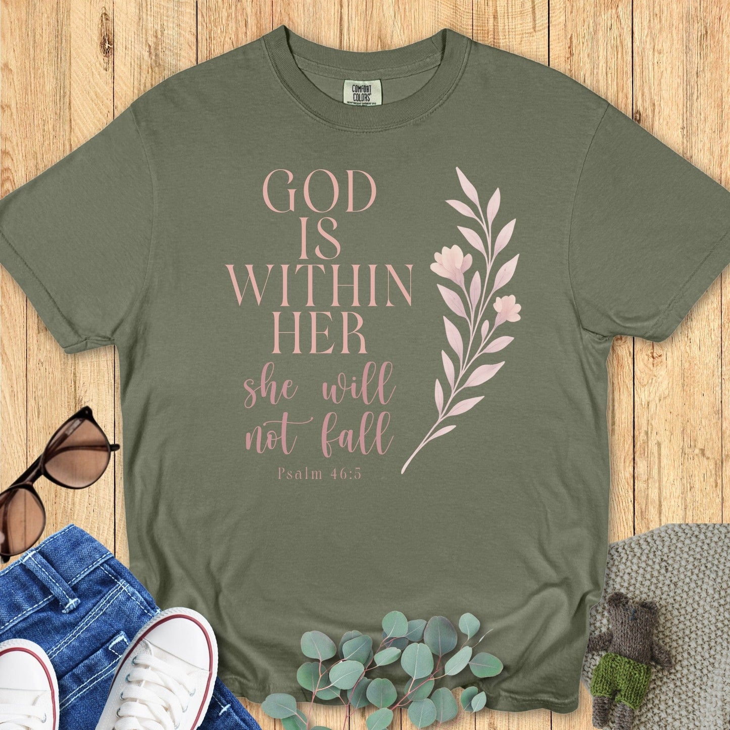 Flatlay of a Comfort Colors 1717 sage “God Is Within Her – Psalm 46:5” t-shirt designed with pastel pink lettering and floral motif. A modern Christian shirt celebrating strength, faith, and grace.