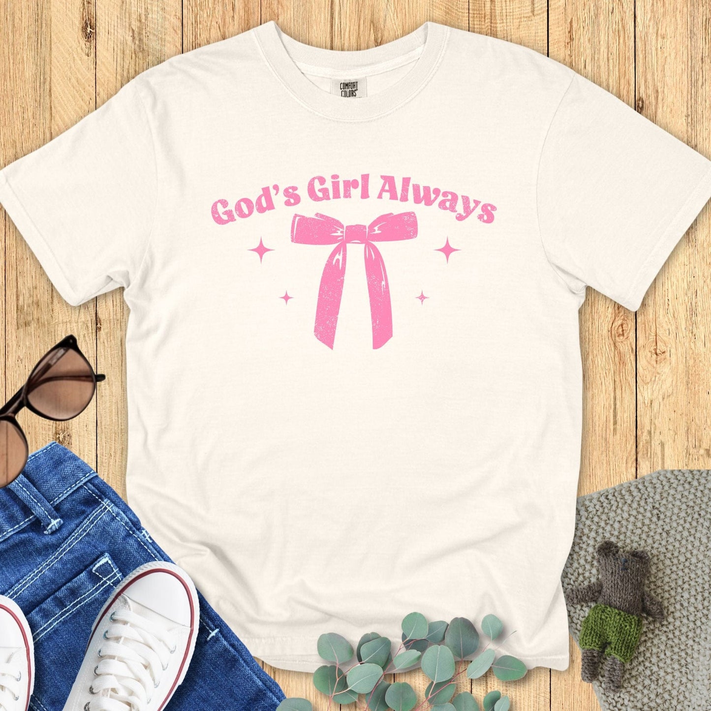 Flat lay of an ivory T-shirt with the same “God’s Girl Always” pink bow and sparkles design, styled with sunglasses, jeans, and greenery.