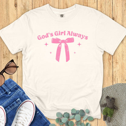 Flat lay of an ivory T-shirt with the same “God’s Girl Always” pink bow and sparkles design, styled with sunglasses, jeans, and greenery.