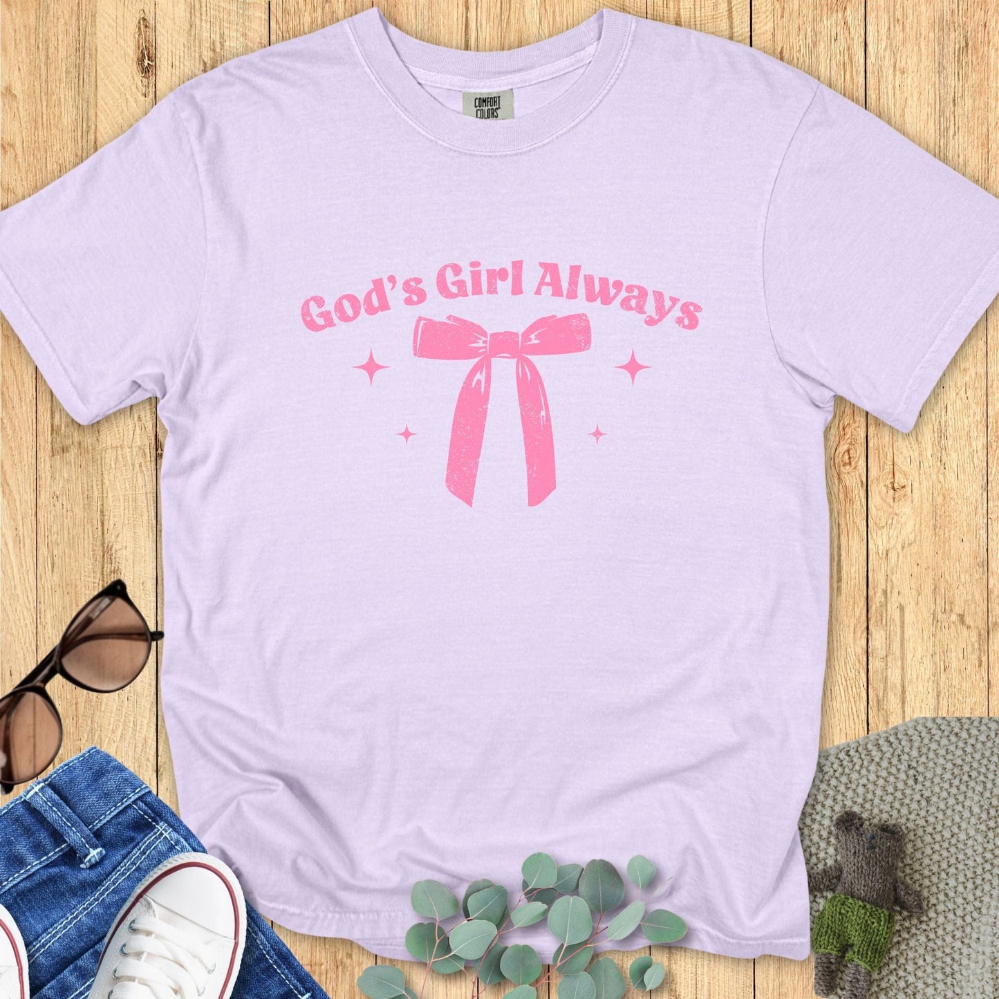 Flat lay of a lavender T-shirt with the phrase “God’s Girl Always” in pink retro font above a large pink bow and small sparkles.