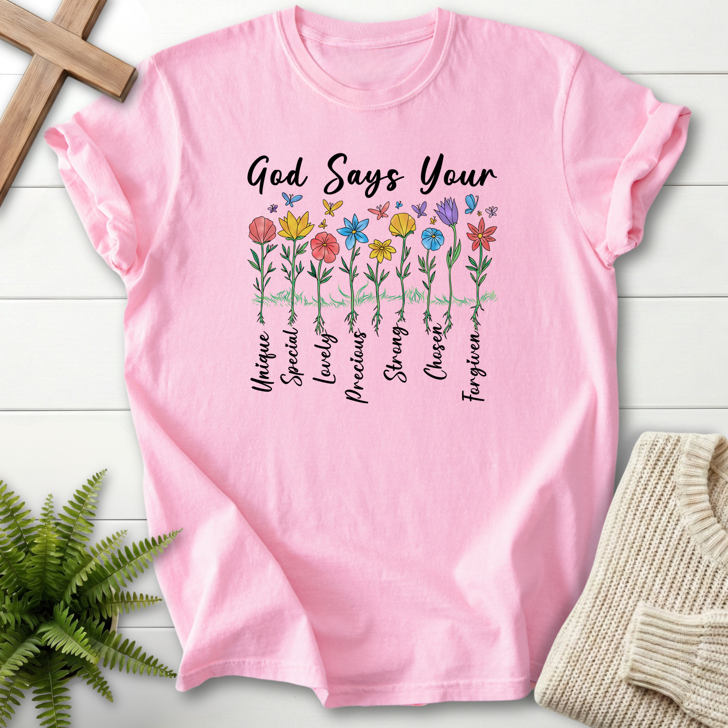 Blossom pink women's faith t-shirt with 'God Says You Are' above a row of wildflowers labeled with affirmations like Chosen and Forgiven.