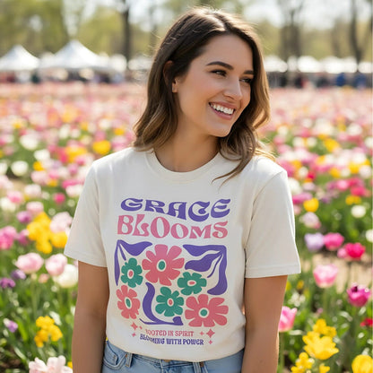 "Young woman smiling in a field of tulips, wearing ivory Comfort Colors 1717 Christian T-shirt with bold retro floral design and text ‘Grace Blooms — Rooted in Spirit, Blooming with Power.’ Vibrant Christian apparel lifestyle photography."