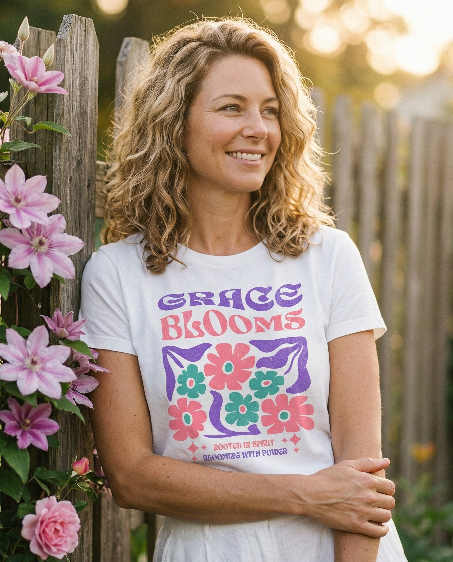Grace Blooms Christian – White women's Christian tee, lifestyle photo in outdoor garden – Retro groovy-style 'GRACE BLOOMS' in bold purple and pink lettering wi – She Wears Faith
