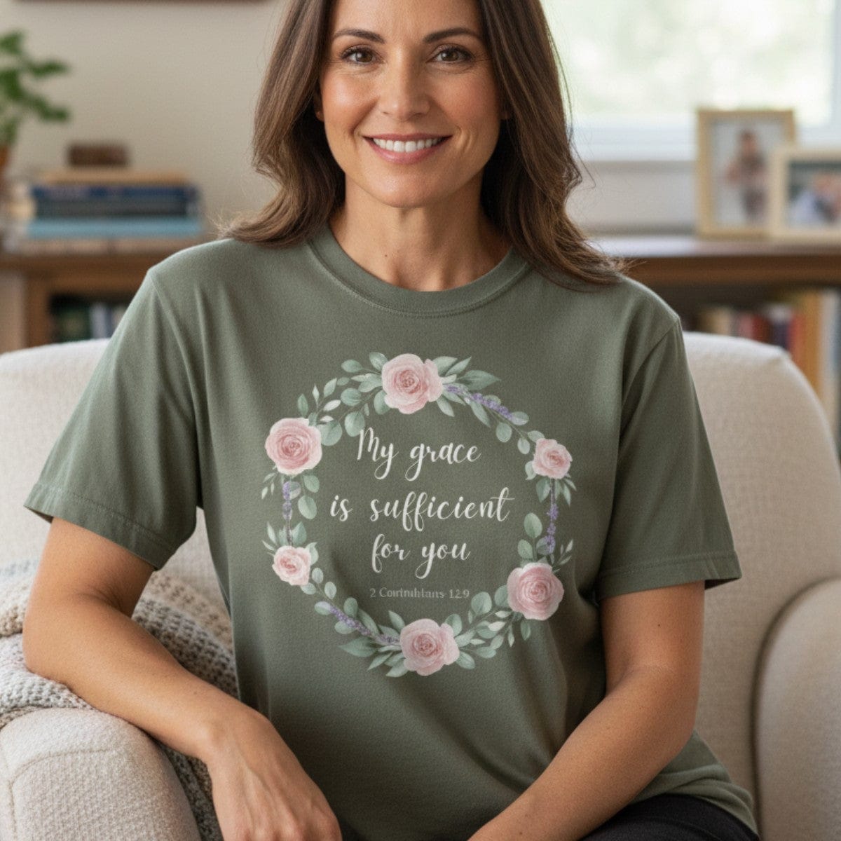 Smiling woman sitting in a cozy living room wearing a Comfort Colors 1717 sage “My Grace Is Sufficient for You – 2 Corinthians 12:9” t-shirt. A soft and inspiring women’s scripture tee, perfect as a Christian mom gift idea.