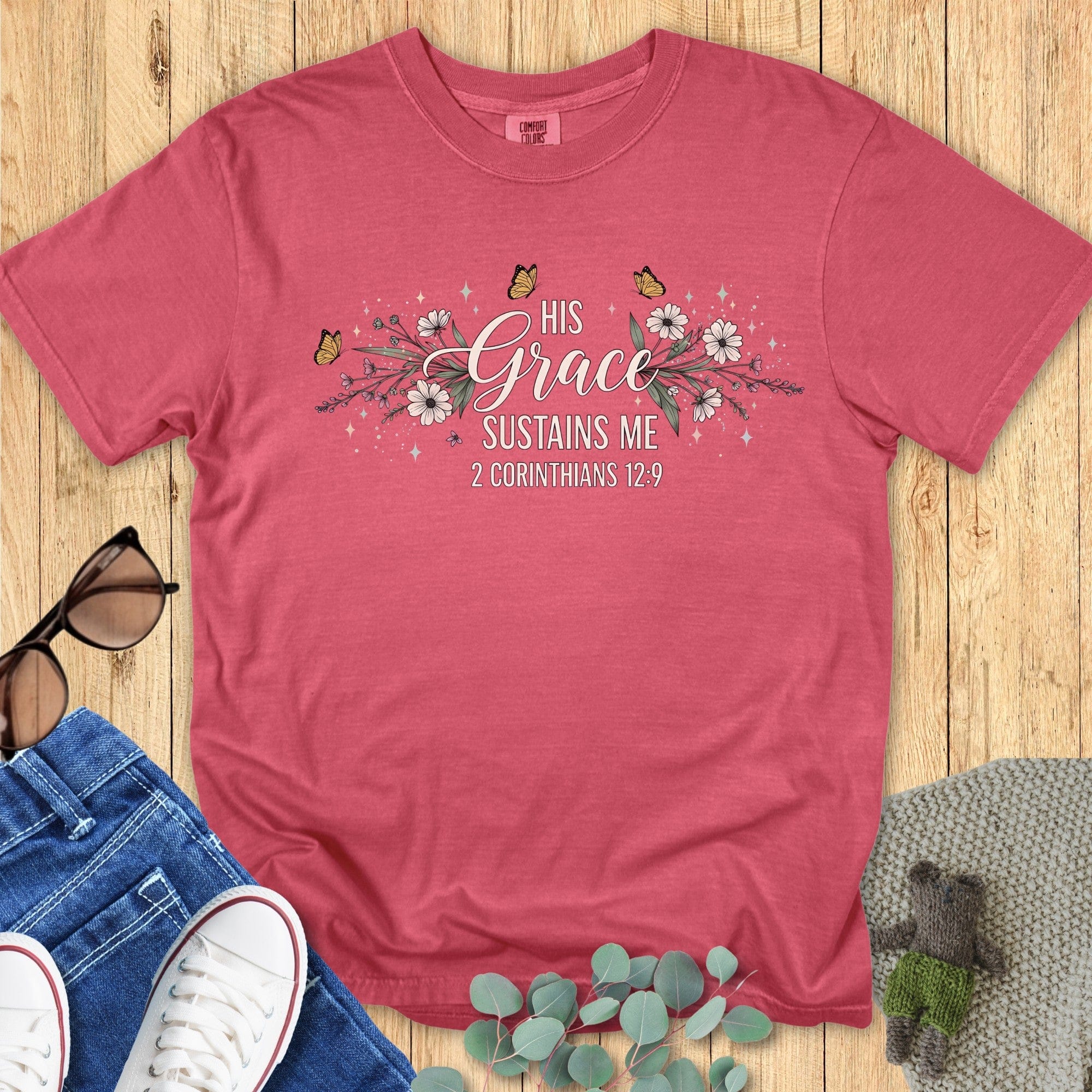 Flatlay of a Comfort Colors 1717 crimson “His Grace Sustains Me – 2 Corinthians 12:9” t-shirt decorated with elegant blossoms and butterflies. A meaningful piece of faith-based apparel for women — ideal Christian mom gift idea or daily encouragement tee.