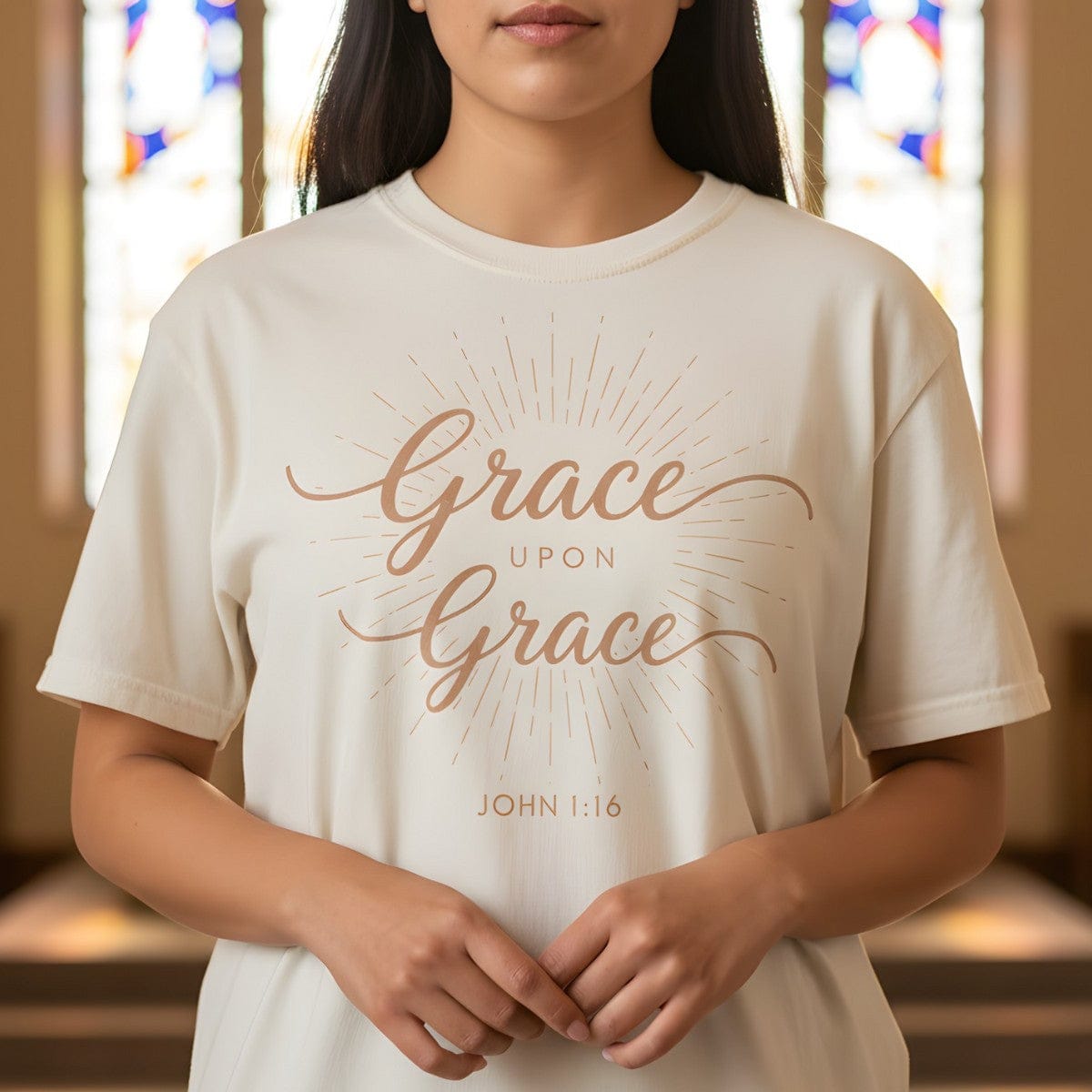 Woman standing inside a softly lit church wearing a Comfort Colors 1717 ivory “Grace Upon Grace – John 1:16” t-shirt with radiant sunburst design. A timeless women’s scripture tee — elegant faith-based apparel for women and modern Christian shirt, meaningful Christian mom gift idea.