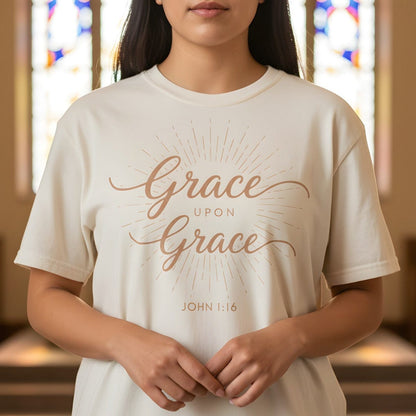 Woman standing inside a softly lit church wearing a Comfort Colors 1717 ivory “Grace Upon Grace – John 1:16” t-shirt with radiant sunburst design. A timeless women’s scripture tee — elegant faith-based apparel for women and modern Christian shirt, meaningful Christian mom gift idea.