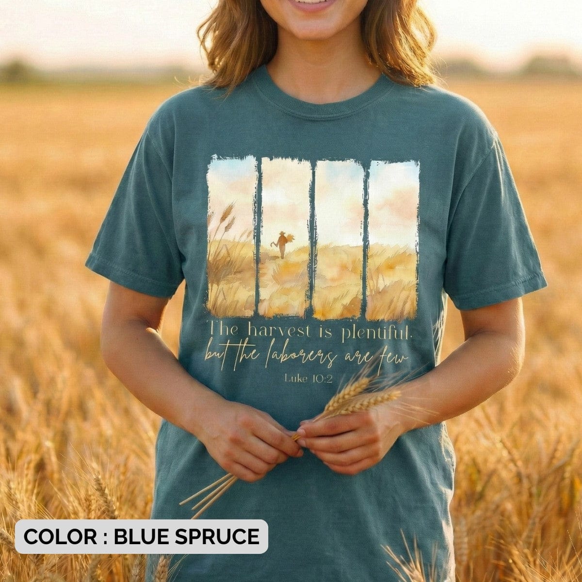 Woman in a Comfort Colors 1717 blue spruce Christian women’s T-shirt with a soft watercolor harvest landscape and the scripture “The harvest is plentiful, but the labourers are few – Luke 10:2.” A serene outdoor US faith wear scene in a wheat field.