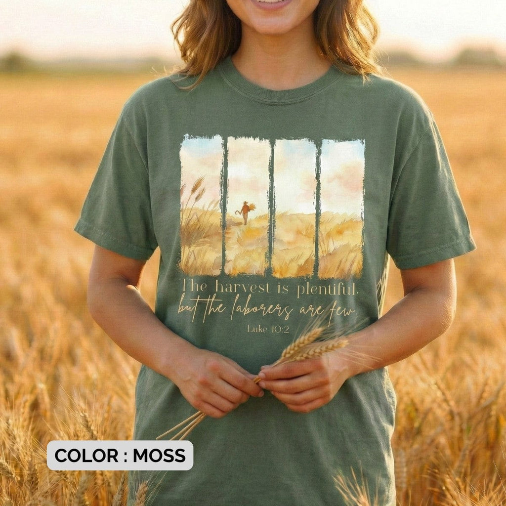 Woman wearing a Comfort Colors 1717 moss Christian women tee showing a watercolor harvest-field illustration and the verse “The harvest is plentiful, but the labourers are few – Luke 10:2.” Natural, rustic US faith wear photographed in a sunlit wheat field.
