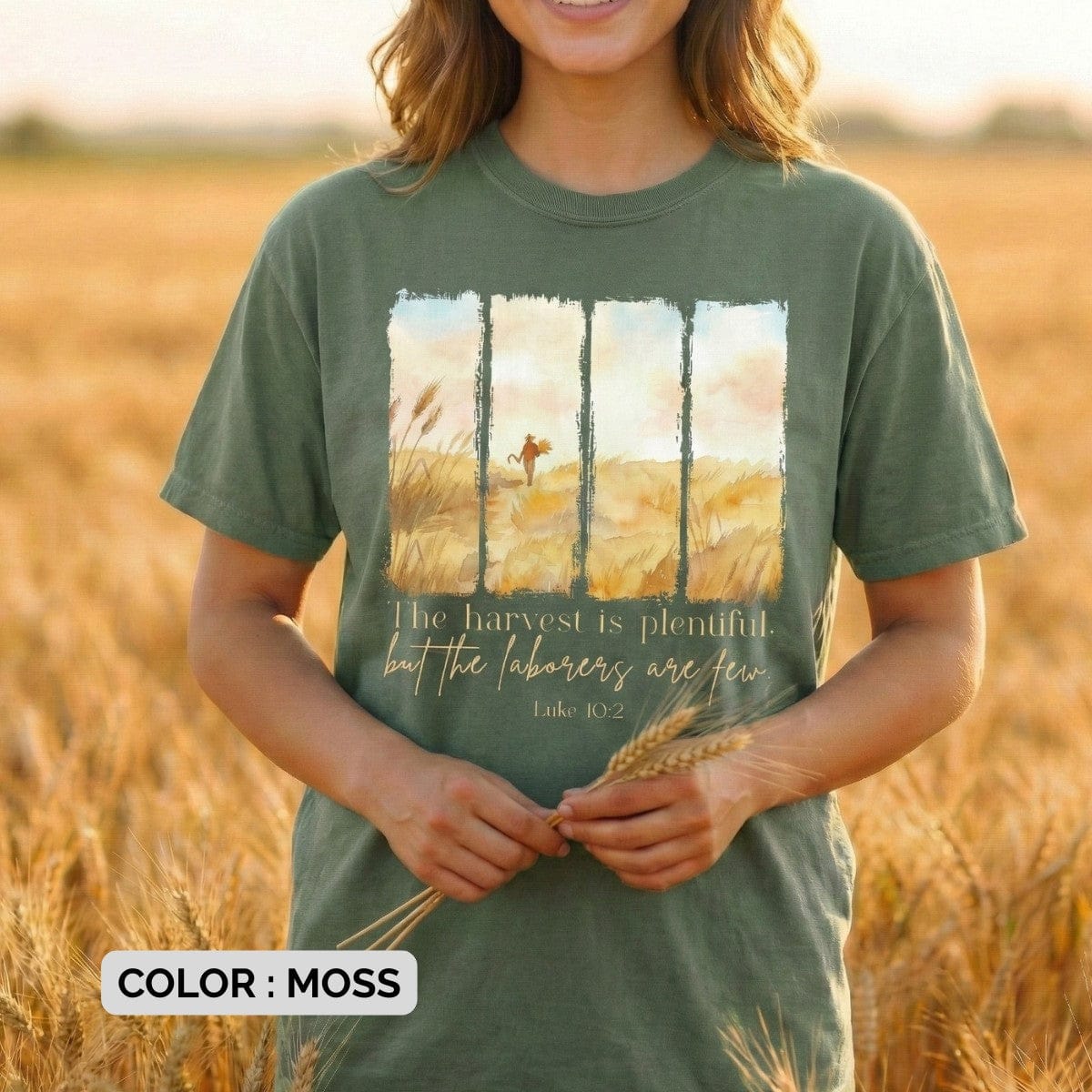 Woman wearing a Comfort Colors 1717 moss Christian women tee showing a watercolor harvest-field illustration and the verse “The harvest is plentiful, but the labourers are few – Luke 10:2.” Natural, rustic US faith wear photographed in a sunlit wheat field.
