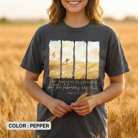 Woman wearing a Comfort Colors 1717 pepper Christian women tee featuring a watercolor harvest-field design with the verse “The harvest is plentiful, but the labourers are few – Luke 10:2.” Styled outdoors in a golden wheat field for a warm US faith wear look.