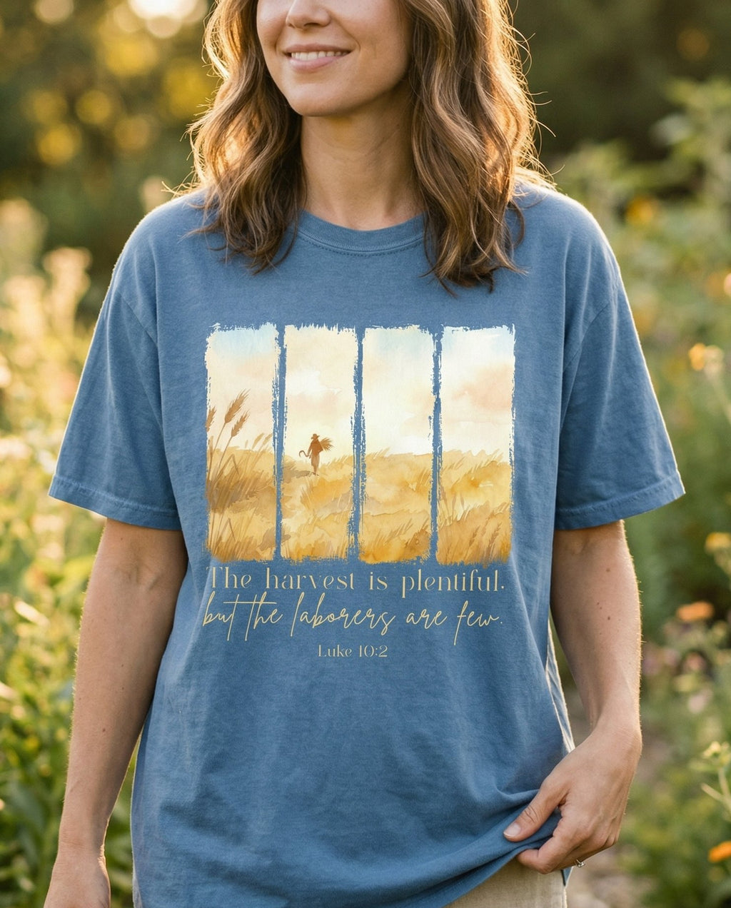Harvest Fields – Blue Jean women's Christian tee, lifestyle photo in outdoor garden – Four watercolor panel illustration of a golden wheat field with a figu – Luke 10:2 – She Wears Faith
