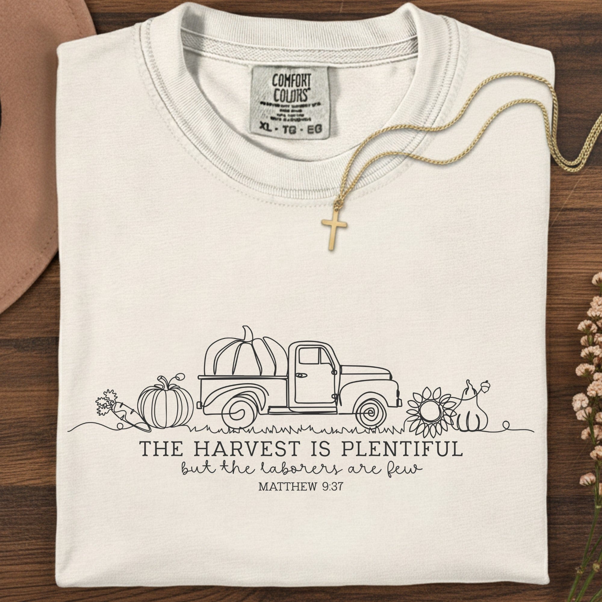 Folded Comfort Colors 1717 ivory “The harvest is plentiful but the laborers are few” t-shirt (Matthew 9:37) styled with gold cross necklace. A neutral women’s scripture tee -  faith-based apparel for women and modern Christian shirt, timeless Christian mom gift idea for the season.