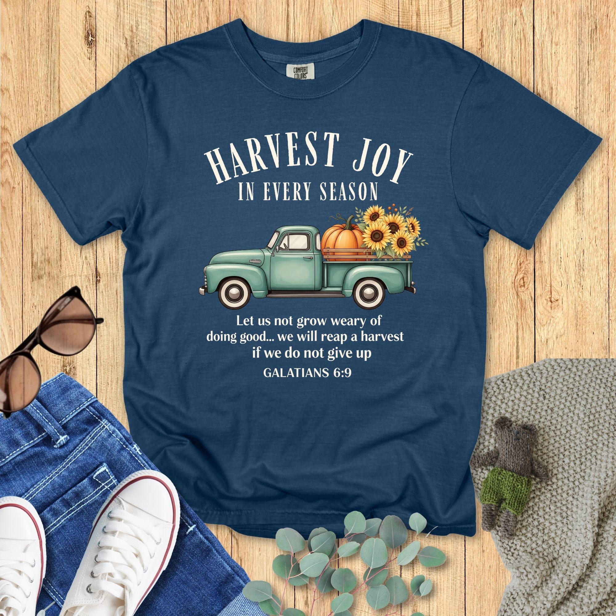 Midnight navy Comfort Colors Christian T-shirt with teal vintage truck, pumpkin, and sunflower harvest graphic. Features scripture "Harvest Joy in Every Season – Galatians 6:9." Inspirational fall faith tee flat lay with jeans and sneakers.