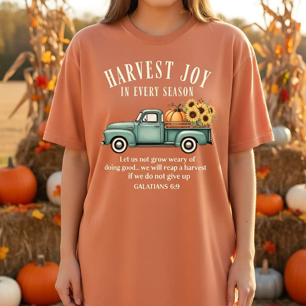 Yam orange Comfort Colors Christian T-shirt with teal vintage truck carrying pumpkins and sunflowers, verse "Harvest Joy in Every Season – Galatians 6:9." Modeled outdoors at a pumpkin patch for a seasonal lifestyle look.