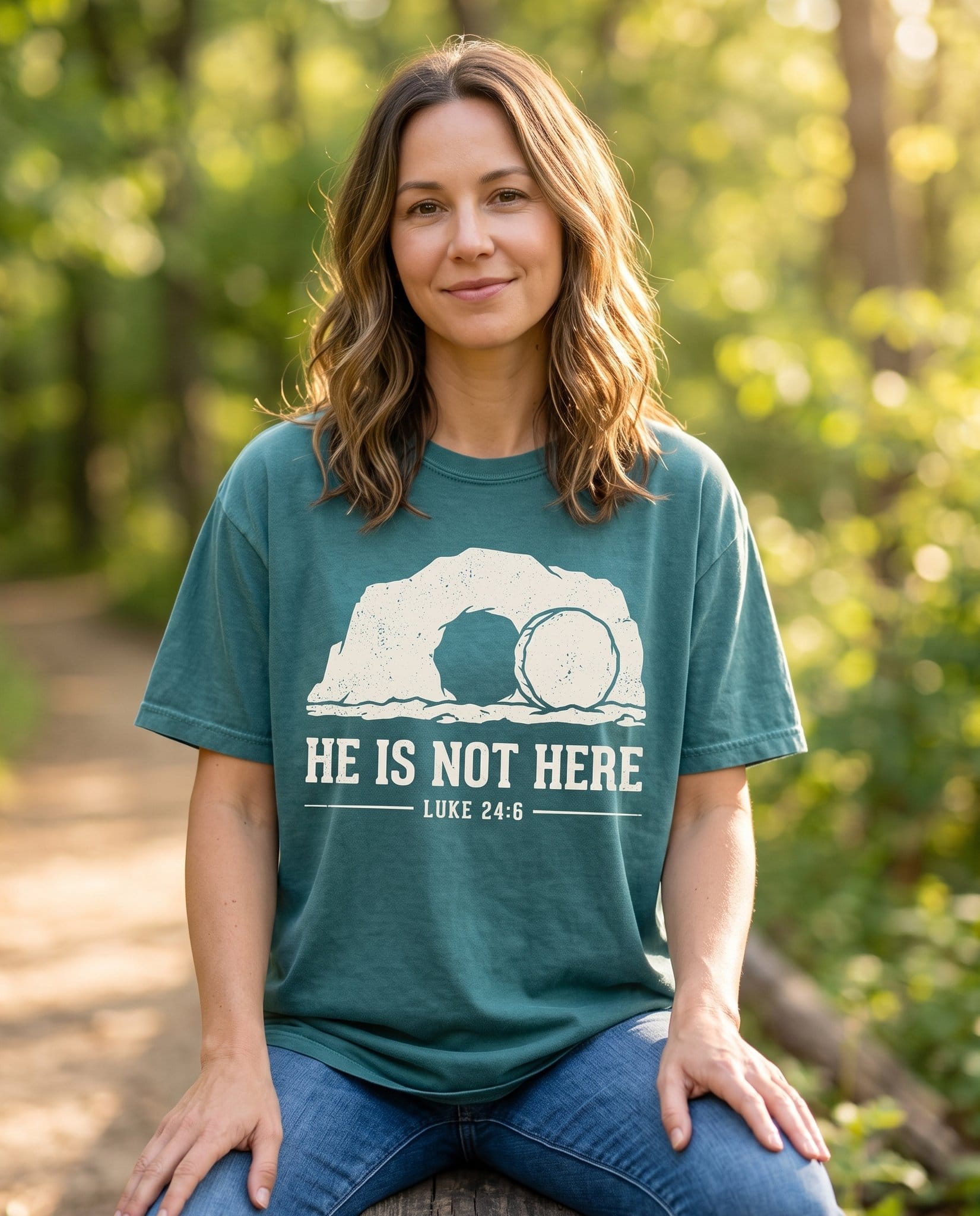 He Is Not Here – Blue Spruce women's Christian tee, lifestyle photo in nature trail – A cream illustration of an open rock tomb with the stone rolled away – Luke 24:6 – She Wears Faith