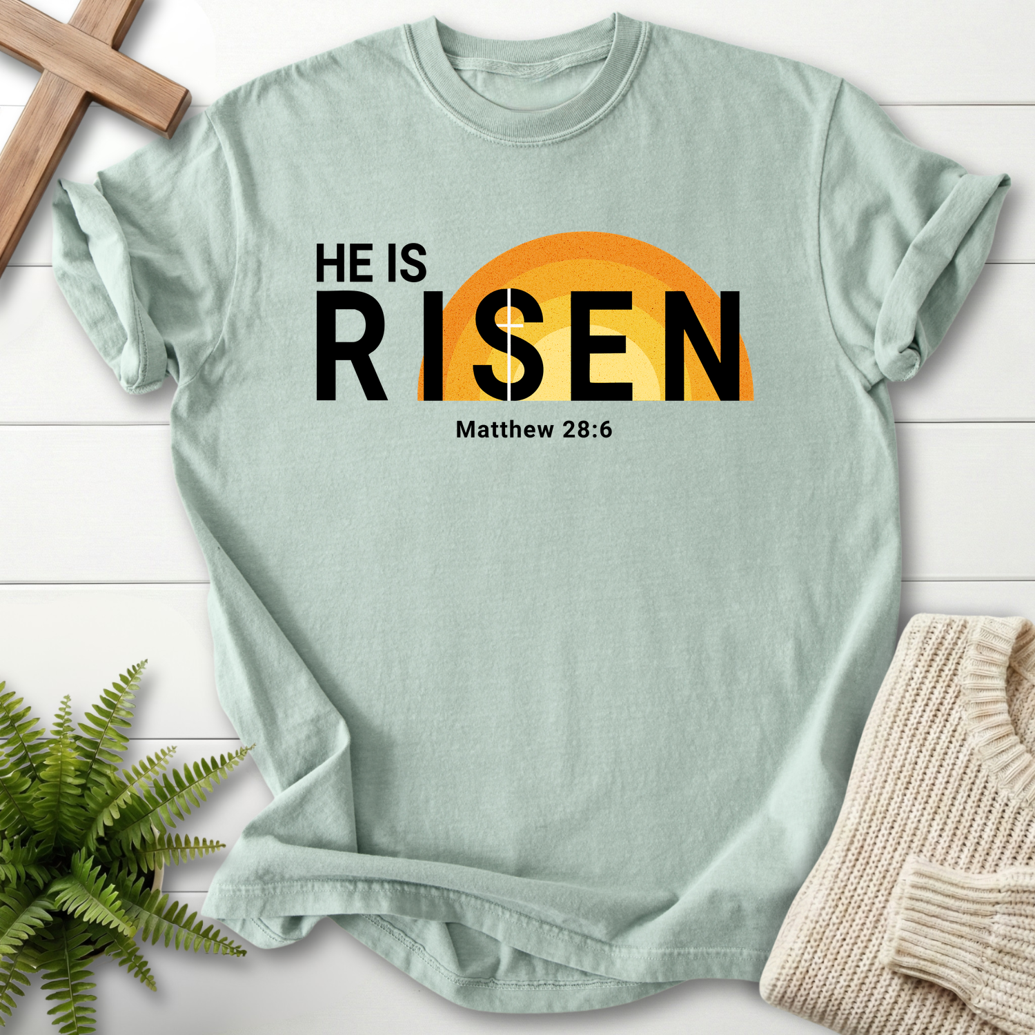 Bay Bible verse tee with bold 'He Is Risen' Easter design and an orange sun rising graphic, inspired by Matthew 28:6.