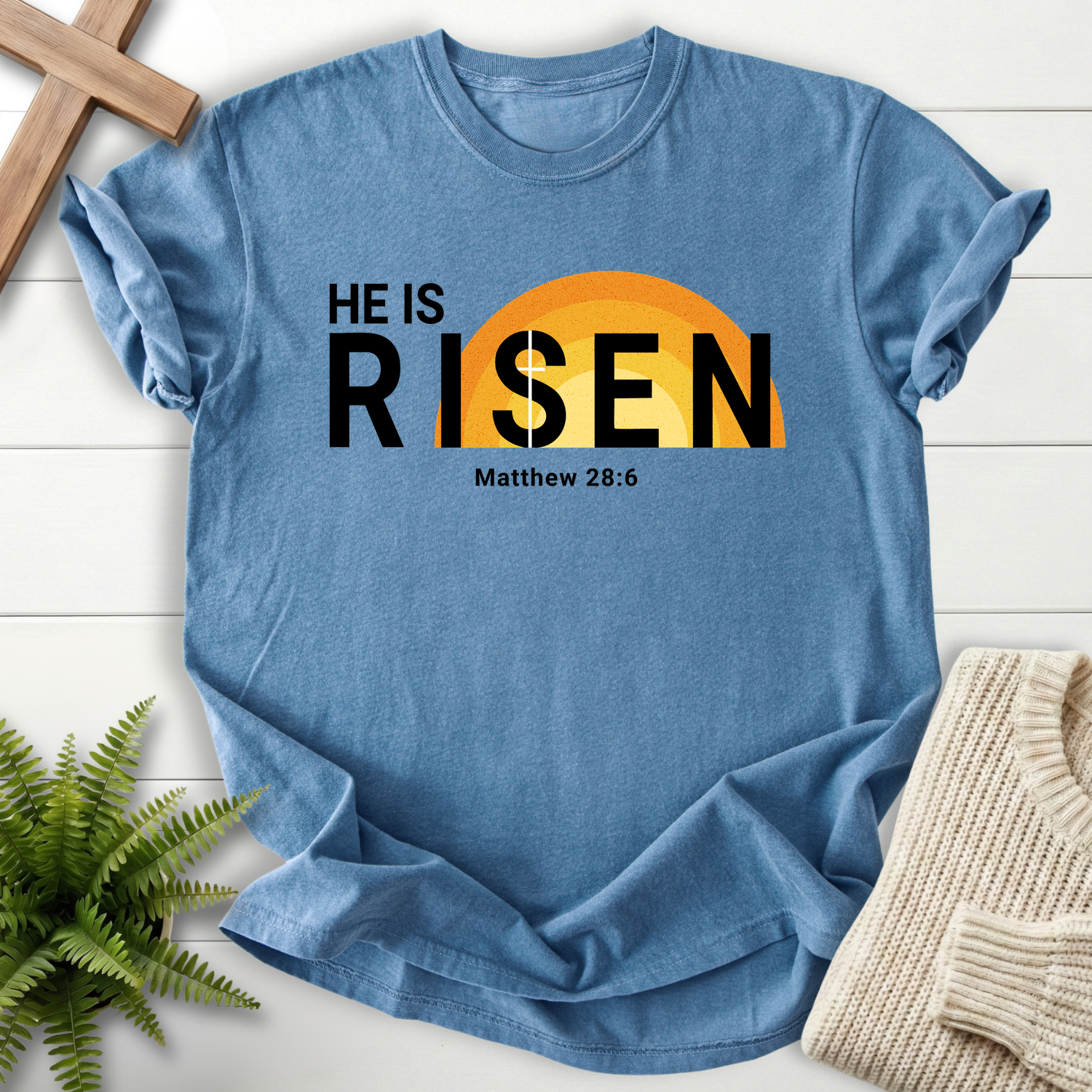 Blue Jean women's faith t-shirt with bold 'He Is Risen' Easter text and a sunrise illustration, inspired by Matthew 28:6.