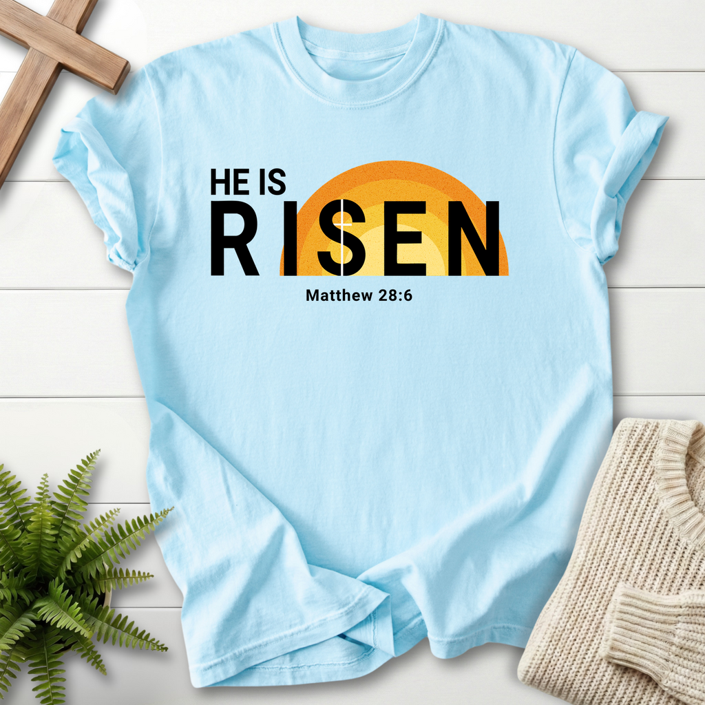 Chambray women's faith t-shirt with 'He Is Risen' in modern bold type and a golden sunrise, inspired by Matthew 28:6.