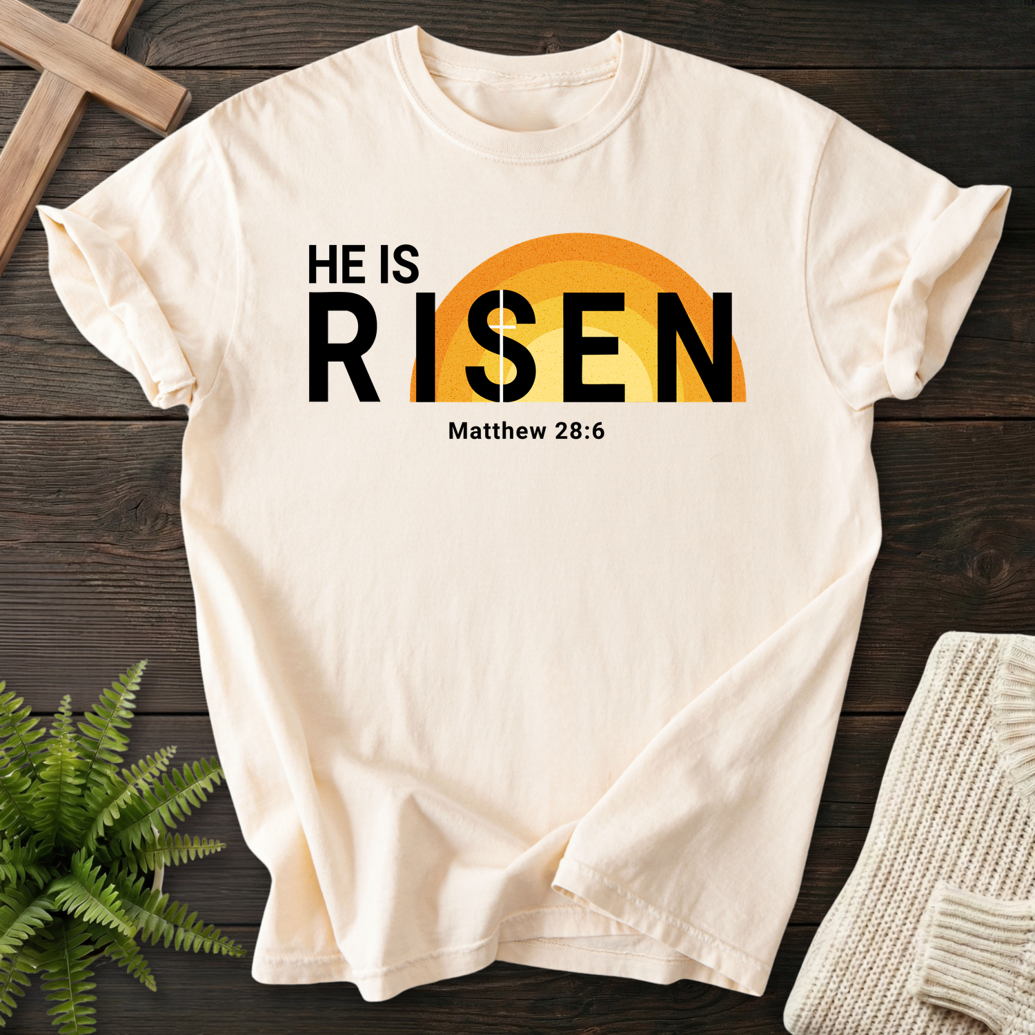 Ivory Bible verse tee with bold 'He Is Risen' lettering and a golden sunrise graphic, inspired by Matthew 28:6.
