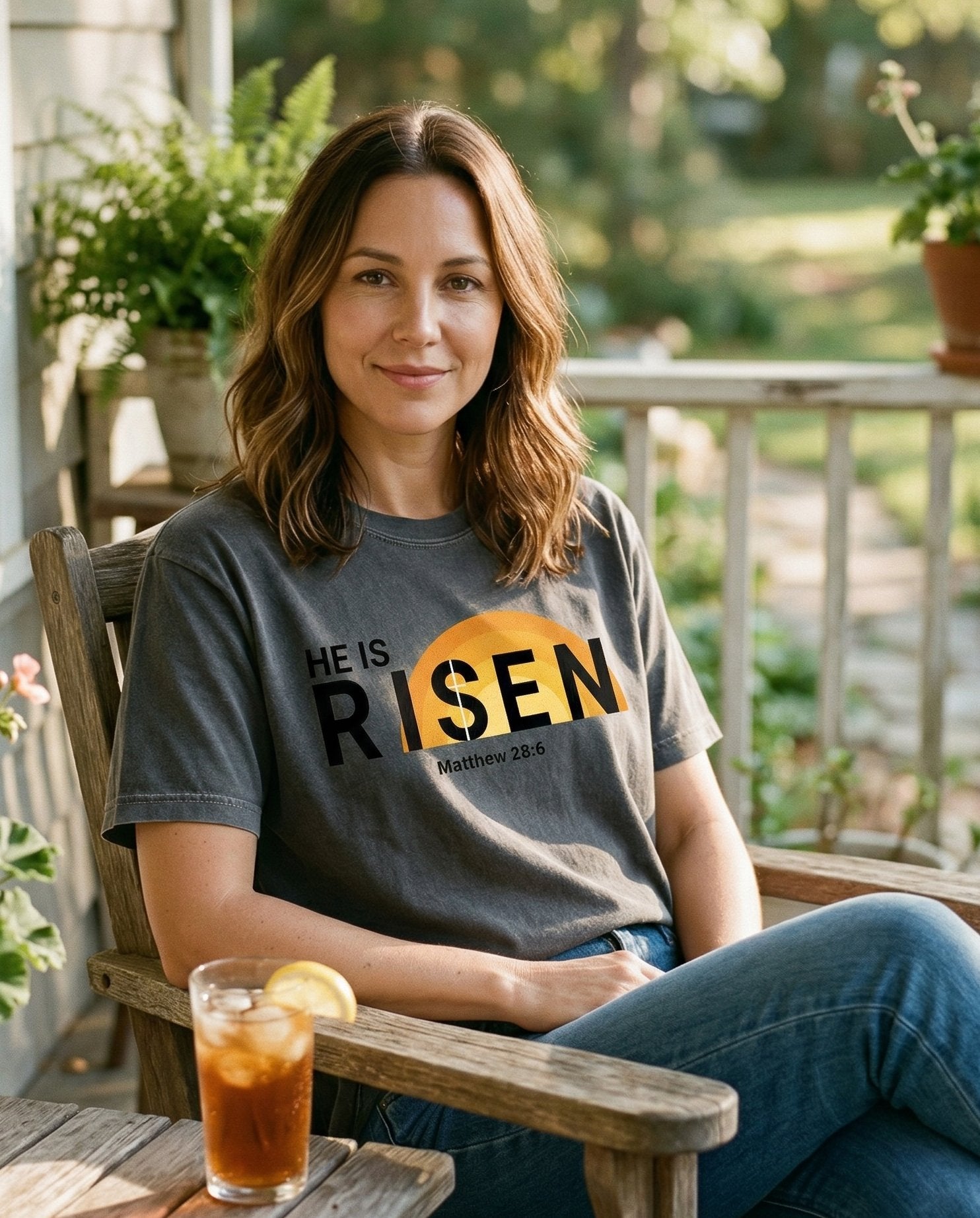 He Is Risen – Pepper women's Christian tee, lifestyle photo in sunny porch – Bold modern typography 'HE IS RISEN' with a large rising sun in orange – Matthew 28:6 – She Wears Faith