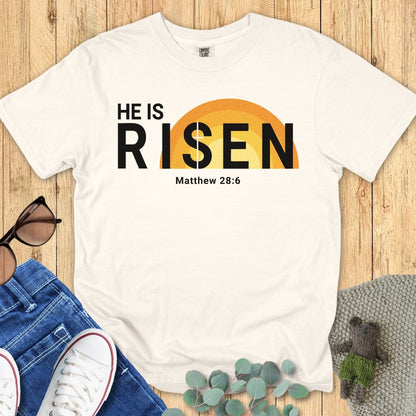 Flatlay of a Comfort Colors 1717 ivory “He Is Risen – Matthew 28:6” t-shirt featuring a bold sunrise graphic and minimalist typography. A bright women’s scripture tee — faith-based apparel for women and modern Christian shirt, thoughtful Christian mom gift idea for Easter.