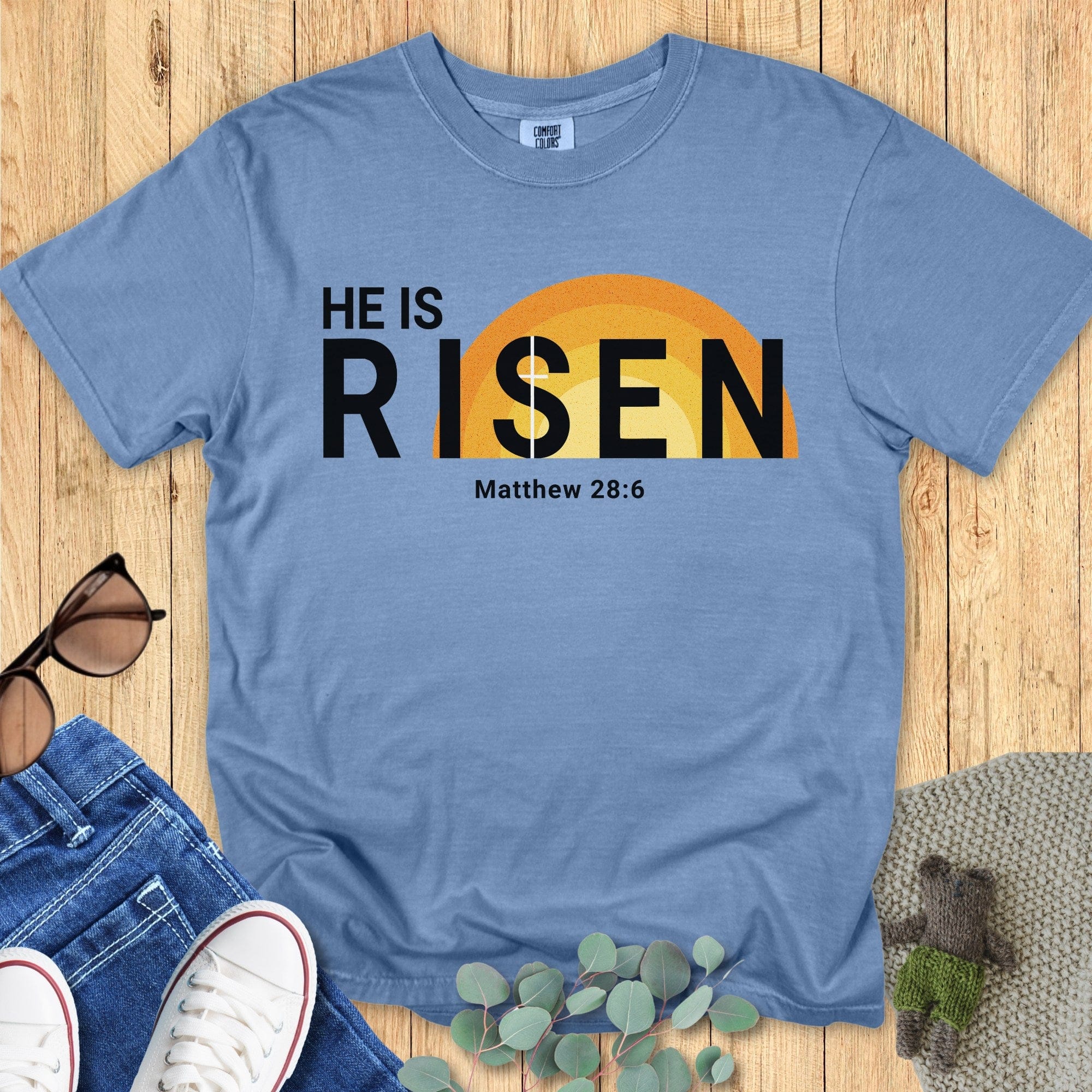 Flatlay of a Comfort Colors 1717 blue jean “He Is Risen – Matthew 28:6” t-shirt showcasing a warm yellow sunrise design. A classic women’s scripture tee — encouraging faith-based apparel for women and modern Christian shirt, inspiring Christian mom gift idea for Easter.