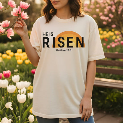 Woman standing in a garden of tulips wearing a Comfort Colors 1717 ivory “He Is Risen – Matthew 28:6” t-shirt with a sunrise design. A joyful women’s scripture tee — uplifting faith-based apparel for women and modern Christian shirt, perfect Easter or Christian mom gift idea.
