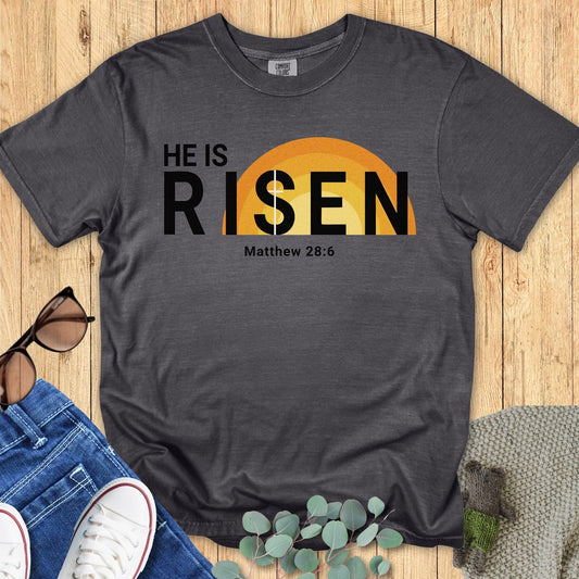 Flatlay of a Comfort Colors 1717 pepper “He Is Risen – Matthew 28:6” t-shirt with a vintage-style sunrise behind bold text. A statement women’s scripture tee — modern faith-based apparel for women and Christian shirt, meaningful Christian mom gift idea.