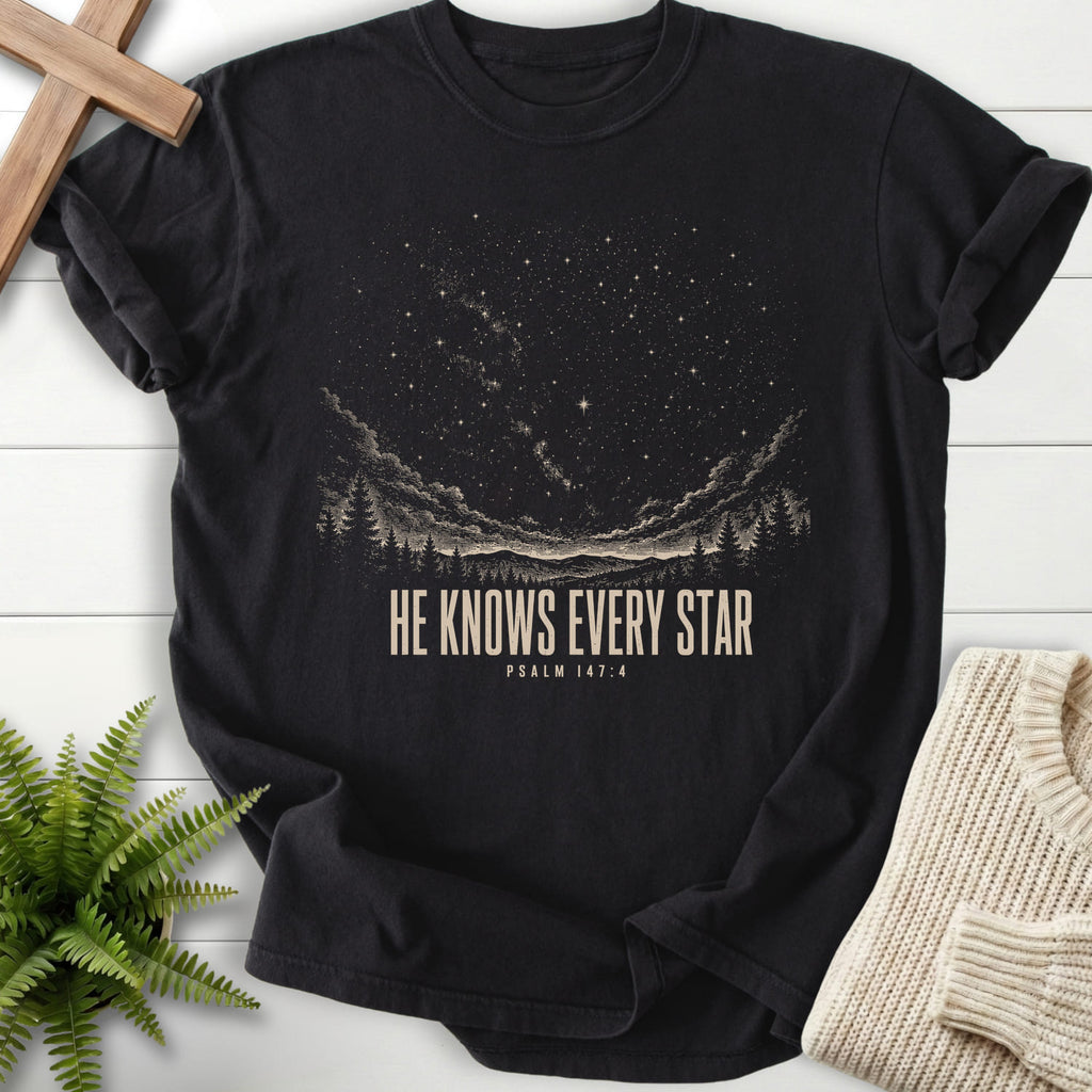 Flat lay of a Black Christian graphic tee featuring mountain graphic with "He Knows Every Star T-Shirt" Psalm 147:4.