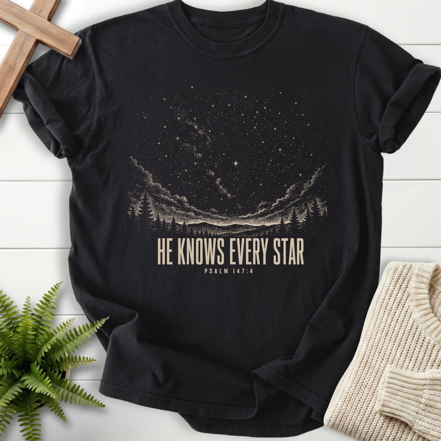 Flat lay of a Black Christian graphic tee featuring mountain graphic with "He Knows Every Star T-Shirt" Psalm 147:4.