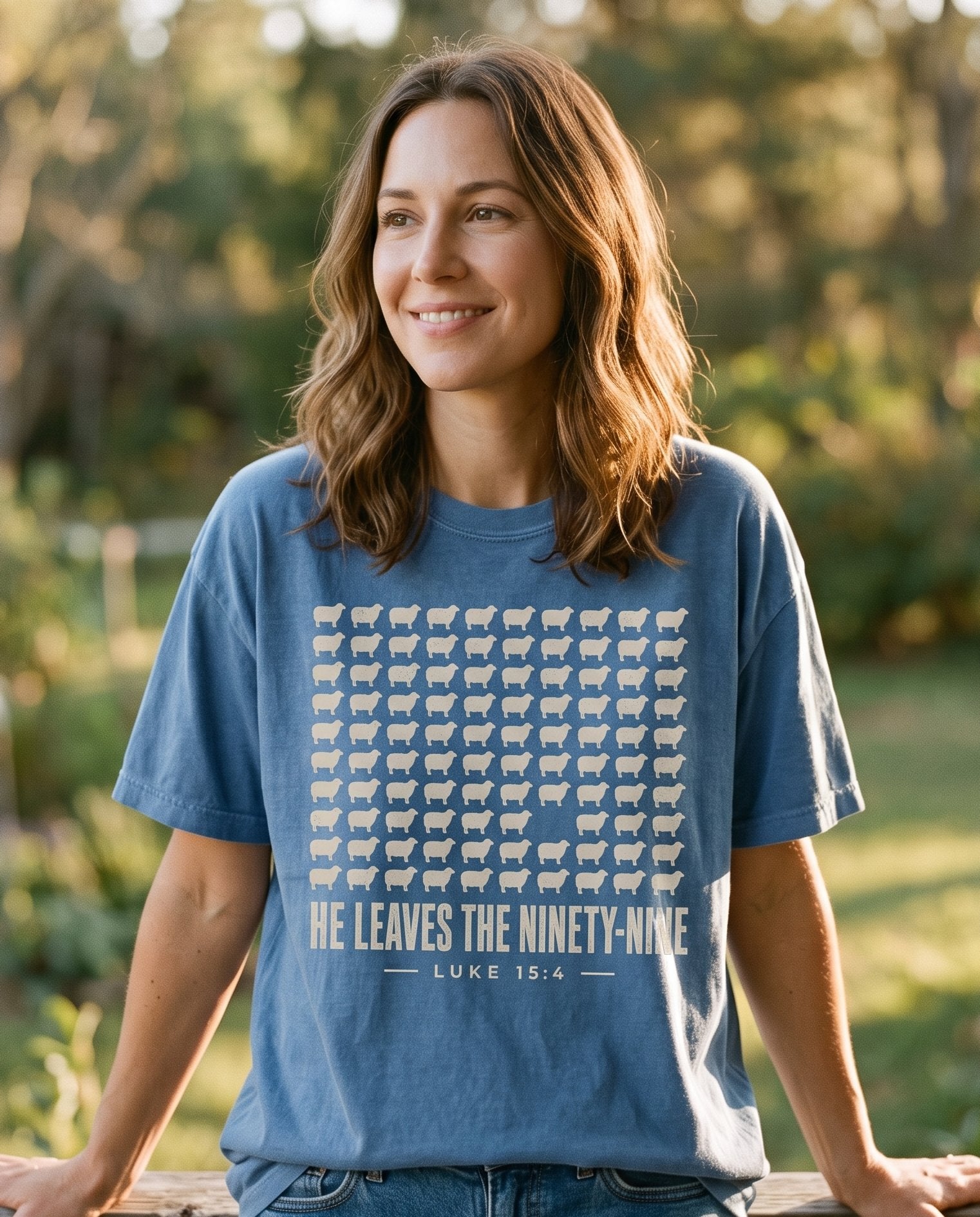 He Leaves the Ninety-Nine – Blue Jean women's Christian tee, lifestyle photo in sunny porch – A grid of ninety-nine tan sheep silhouettes with one sheep missing fro – Luke 15:4 – She Wears Faith
