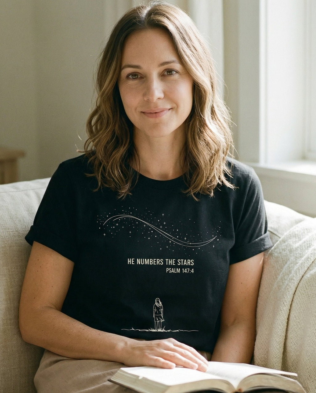 He Numbers the Stars – Black women's Christian tee, lifestyle photo in cozy living room – Line-art illustration of a woman standing beneath a sweeping Milky Way – Psalm 147:4 – She Wears Faith