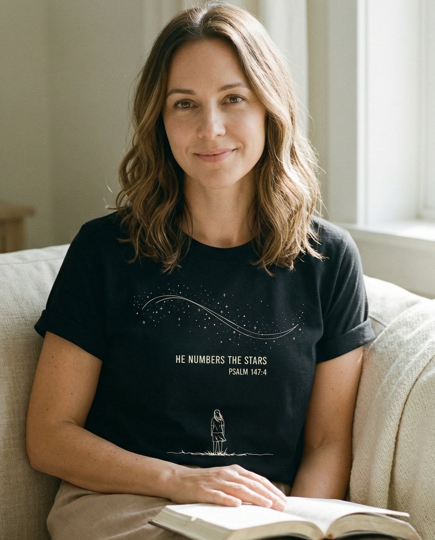 He Numbers the Stars – Black women's Christian tee, lifestyle photo in cozy living room – Line-art illustration of a woman standing beneath a sweeping Milky Way – Psalm 147:4 – She Wears Faith