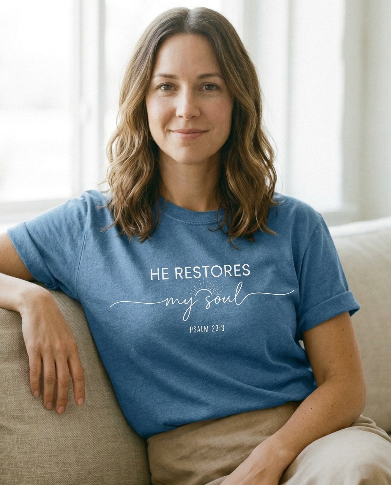 He Restores My Soul T-shirt – Blue Jean women's Christian tee, lifestyle photo in cozy living room – Bold block lettering 'HE RESTORES' above a small sunburst illustration – Psalm 23:3 – She Wears Faith